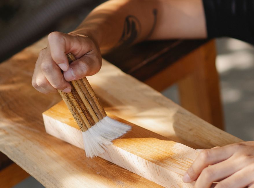 High angle of unrecognizable artisan using brush for varnishing wooden beam while working in professional workshop